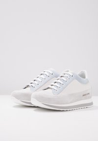 Vita och ljusblå sneakers med en överdel i mocka och läder, platta skosnören och en dämpad sula med ett strukturerat mönster. Varumärkesdetalj synlig.