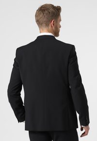 Giacca nera su misura con una texture liscia, un'apertura singola sul retro e bottoni lucidi sui polsini. Colletto della camicia bianca visibile.