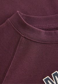 Bordeaux sweatshirtstof med en struktureret overflade. Har en rund halsudskæring og synlig syninger. Indeholder et grafisk logo i kontrasterende farver.