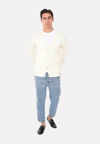 Cardigan color crema con scollo a V, chiusura a bottoni e orlo a costine, indossato sopra una maglietta bianca, abbinato a jeans azzurri chiari e scarpe nere.