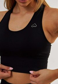 Czarny stanik sportowy z okrągłym dekoltem, z logo serca w białym kolorze po lewej stronie oraz teksturowaną elastyczną taśmą z logo na dole.