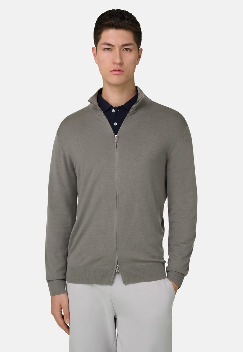 Cardigan gris clair à fermeture éclair avec un col montant et des poignets côtelés, fabriqué en tissu tricoté lisse. Porté par-dessus un polo bleu marine.