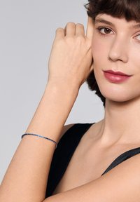 Srebrna bransoletka ozdobiona niebieskimi i przezroczystymi kamieniami, o smukłym i cienkim designie. Noszona na nagim nadgarstku, na tle czarnej bluzki.