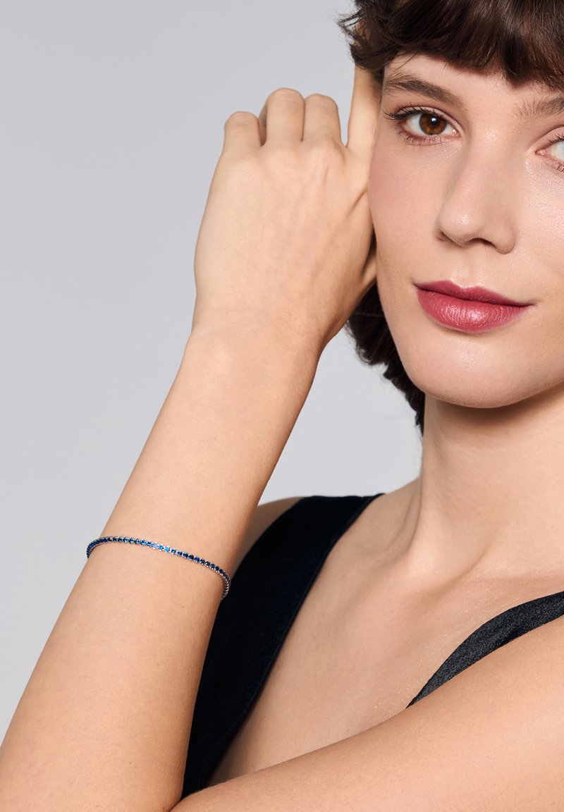 Srebrna bransoletka ozdobiona niebieskimi i przezroczystymi kamieniami, o smukłym i cienkim designie. Noszona na nagim nadgarstku, na tle czarnej bluzki.