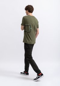 T-shirt vert olive à manches courtes avec un design de poche rectangulaire imprimée ; associé à un pantalon noir et des baskets noires avec des semelles blanches.