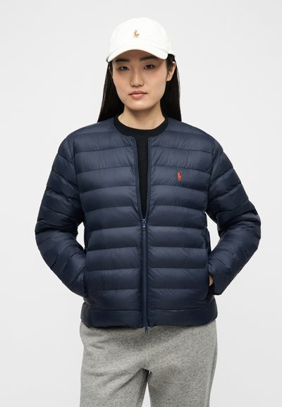 Γυναίκα που φοράει σκοτεινόμπλε μπουφάν με κόκκινο λογότυπο, γκρι παντελόνι και λευκό καπέλο του μπέιζμπολ, στέκεται με τα χέρια στις τσέπες σε απλό φόντο.