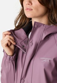 Paarse waterdichte jas met een hoge kraag, drukknoopsluitingen en een logo. Zachte textuur, elastische manchetten en ritszakdetails.