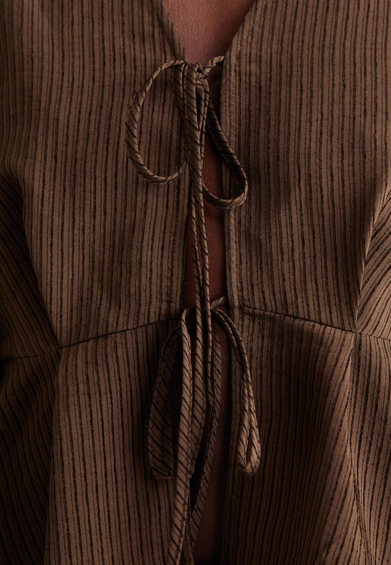 Brązowa, paskowana bluzka z dekoltem w kształcie litery V, z przodu z wiązaniem i luźnym krojem. Tekstura wydaje się miękka i lekko opadająca.