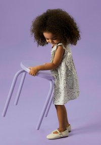 Violetti metallinen jakkara, jossa on pyöristetty istuin ja jalat, jota lapsi pitää kukkamekossa. Sileä pinta pehmeää purppuraista taustaa vasten.