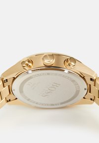 Montre-bracelet dorée avec un bracelet en métal élégant, trois boutons-poussoirs et un boîtier rond. Présente un fond transparent et un texte gravé.