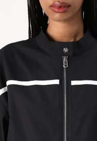 Black jacket with white horizontal stripes, featuring a high collar and silver zipper with a snap closure. Smooth, lightweight fabric.