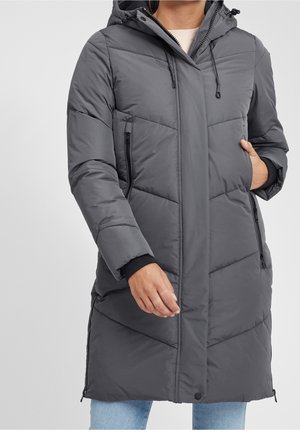 Personne portant un long manteau d'hiver gris matelassé avec capuche, poches zippées et fermetures éclair latérales, habillée d'un jean bleu clair.