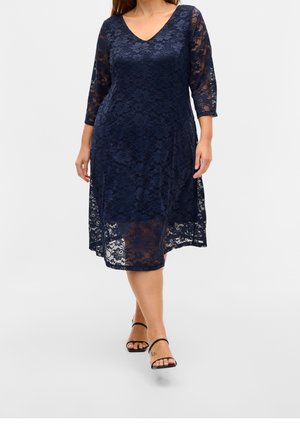 Kobieta idzie, ubrana w granatową koronkową sukienkę do kolan z rękawami do łokci oraz czarne sandały na paski.