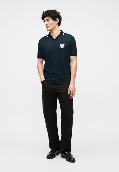 Νέος άνδρας με σγουρά μαλλιά, που φορά μαύρο πόλο με ροζ έμβλημα, μαύρο παντελόνι και μαύρα δερμάτινα παπούτσια, στέκεται με το ένα χέρι στην τσέπη.