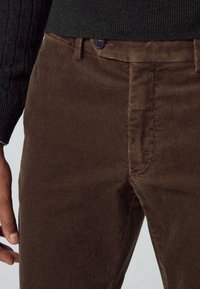 Pantalones de pana marrones con una textura de estría fina, que cuentan con un cierre de botón y bolsillos frontales. Combinados con un suéter de punto verde oscuro.