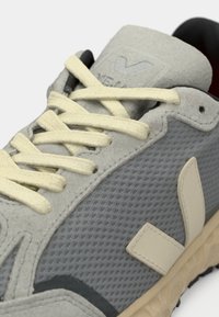 Lähikuva harmaasta lenkkarista, jossa on kermaiset nauhat, verkkokankainen sivu, anteeksi suomukuiduista tehdyt päällysteet ja beige logo yksityiskohtana texturoituneella pohjalla.