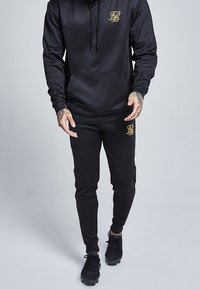 Chándal negro que incluye una sudadera con capucha y un bolsillo frontal, con un logo dorado, combinado con pantalones ajustados. Tejido texturizado y corte atlético.