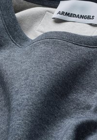 Close-up of folded grey fabric shirt with sewn-in white label showing "ARMEDANGELS" brand name in black text.