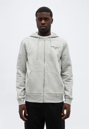 Férfi világosszürke, elöl cipzáros kapucnis pulóverben, elülső zsebekkel és kisebb fekete logóval a mellkasán, egyszínű háttér előtt áll.