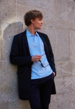 Light blue polo shirt under a black coat, standing against a stone wall. Holding sunglasses, with a relaxed posture. Dark pants complete the outfit.