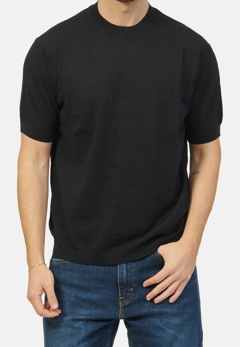 Homme portant un t-shirt en maille noir uni à manches courtes et un jean bleu, une main dans la poche, sur fond uni.