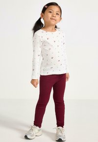 Valkoinen pitkähihainen toppi, jossa on pieniä vaaleanpunaisia kukkia, yhdistettynä viininpunaisiin ribbineulosleggings-housuihin. Lapsella on harmaat lenkkarit, joissa on valkoisia yksityiskohtia.