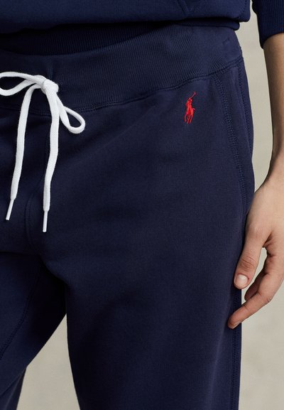 Μπλε σκούρο φούτερ παντελόνι με λευκό κορδόνι, κόκκινο κεντημένο λογότυπο Polo και ένα χέρι να ακουμπάει στον δεξί μηρό.