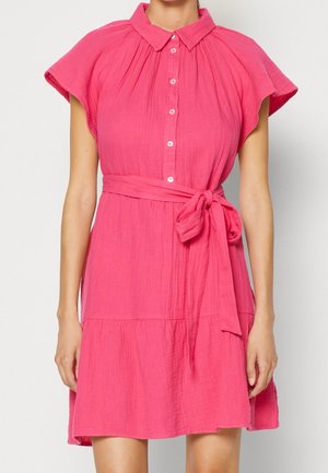 Roze katoenen jurk met een kraag, korte mouwen, knoopsluiting en een ceintuur die in een strik wordt gebonden. Heeft een rok met meerdere lagen.