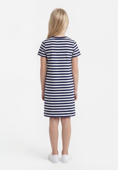 Παιδί με μακριά ξανθά μαλλιά, φορώντας κοντομάνικο φόρεμα με ναυτικό μπλε και άσπρες ρίγες και λευκά αθλητικά παπούτσια, γυρισμένο με την πλάτη σε απλό λευκό φόντο.