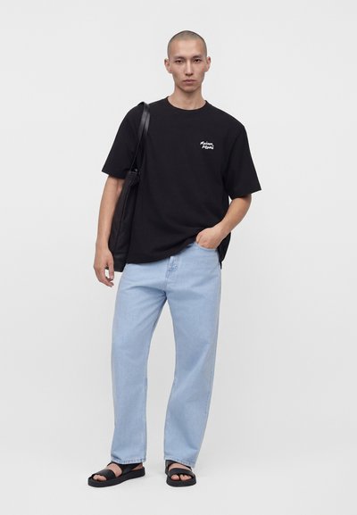 Άνδρας στέκεται, φοράει μαύρο γραφικό t-shirt, ανοιχτό μπλε τζιν, μαύρα σανδάλια και κρατάει μαύρη τσάντα ώμου σε λευκό φόντο.