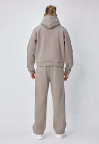 Personne aux cheveux tressés portant un sweat à capuche beige assorti et un pantalon de survêtement, debout face à un fond gris clair uni, avec des baskets blanches visibles.