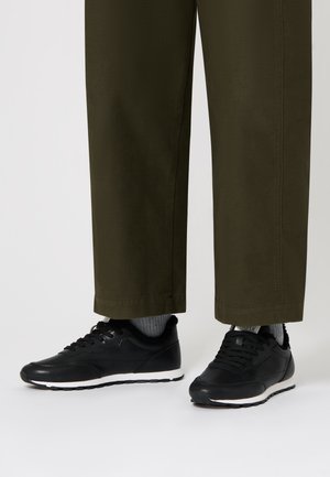 Scarpe da ginnastica nere con tomaia in pelle liscia, suola bianca e lacci, abbinate a pantaloni cropped verde oliva e calzini grigi.