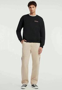 Um jovem vestindo um sweatshirt preto, calças cargo bege e ténis pretos, de pé contra um fundo claro liso.