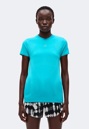 Femme portant un t-shirt de sport bleu vif à manches courtes et un short à motif noir et blanc, debout devant un fond uni.