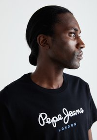 Sort bomulds t-shirt med et hvidt, kursivt logo "Pepe Jeans" og "LONDON" nedenunder i en mindre skrifttype. Model vist i profil.