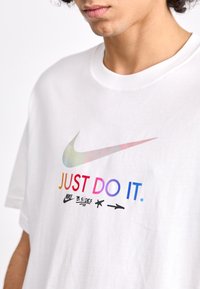 Balta trumpa rankovėmis marškinėliai su spalvingu "Nike" logotipu ir "JUST DO IT." tekstu gradientinėse spalvose, pagaminti iš švelnaus medvilnės audinio.
