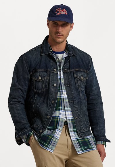 Donkerdenim jack over een groen en blauw geruit shirt, met daaronder een wit T-shirt, aangevuld met een marineblauwe pet met een rood logo.