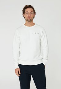 Sudadera blanca de manga larga con cuello y puños de canalé, que presenta un pequeño logo negro de "SAIL RACING" en el pecho. Textura suave.