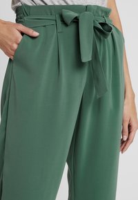Pantaloni verdi a vita alta realizzati in un tessuto liscio, con cintura da annodare e dettagli plissettati sul davanti.