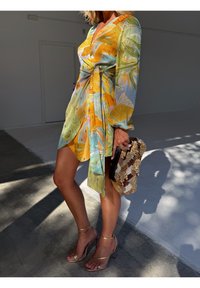 Femme portant une robe portefeuille colorée avec des motifs de feuilles, tenant une pochette perlée et portant des sandales dorées à talons hauts, debout à l'intérieur.