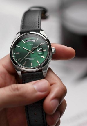 Montre Timex avec un cadran vert, des index argentés, un bracelet en cuir noir, une fonction date et étanche, tenue dans une main.