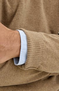 Beżowy wełniany sweter z ribbami przy mankietach, na jasnoniebieskiej koszuli z kołnierzykiem. Tekstura wygląda na miękką, z luźnym dopasowaniem wokół nadgarstka.
