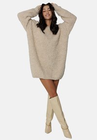 Beżowy, oversize'owy sweter dzianinowy o fakturowanym wykończeniu, z zaokrąglonym dołem i długimi rękawami, zestawiony z beżowymi kozakami do kolan.