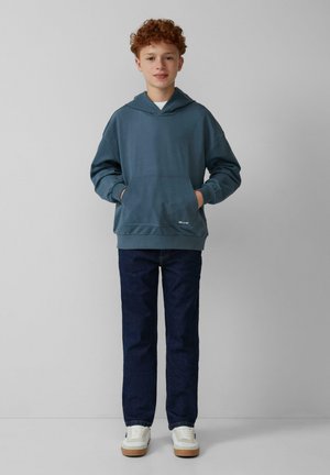 Felpa con cappuccio in teal con tasca frontale, abbinata a jeans scuri e sneakers bianche. Design semplice, vestibilità casual e texture morbida.