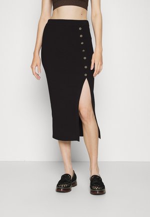 Woman wearing a black midi skirt with side buttons and slit, paired with black patterned loafers and a cropped top.