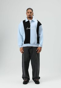 Pull-over à demi-fermeture éclair en mélange de coton bleu clair et noir, avec un grand col et un texte logo. Associé à un pantalon en denim noir ample et des chaussures noires.