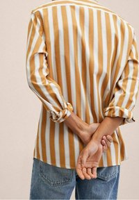 Camicia a righe in cotone giallo senape e bianco, maniche lunghe arrotolate, vista posteriore, con le mani del indossatore intrecciate dietro. Jeans blu casual.