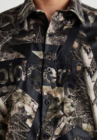 Cămașă cu mânecă scurtă în imprimeu de camuflaj, având o combinație de nuanțe de maro, verde și negru. Butoane la guler și un element de design grafic.