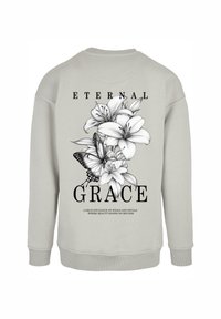Lys grå sweatshirt med et blomsterdesign med liljer og en sommerfugl på ryggen, samt tekst der siger "ETERNAL GRACE."