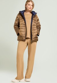 Bronze pufferjakke med navy hætte, der har et quiltet design. Beige langærmet top og tætsiddende bukser, kombineret med hvide sneakers.